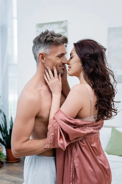 Vista lateral da mulher sorridente em robe de cetim beijando marido sem camisa no quarto — Fotografia de Stock