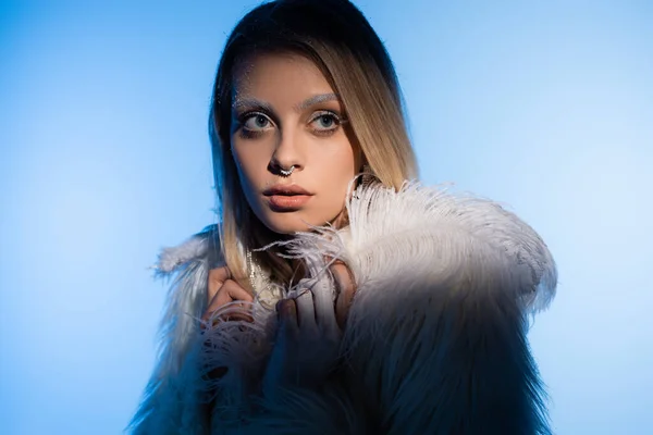 Jeune femme blonde avec des sourcils neigeux en veste avec des plumes regardant loin sur fond bleu — Photo de stock