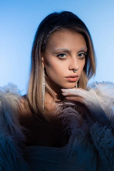 Young blonde woman with snowy eyebrows in jacket with feathers looking at camera on blue — Stock Photo