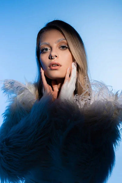 Femme blonde avec des sourcils neigeux en veste avec des plumes regardant la caméra sur bleu — Photo de stock