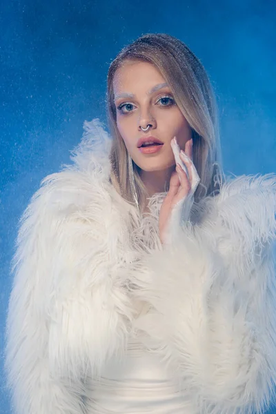 Young pierced woman with snow on hair and winter makeup looking at camera under falling snow on dark blue — Stock Photo