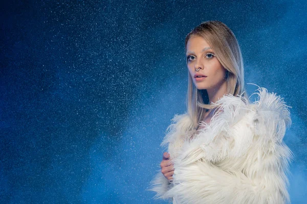 Young pierced woman with snow on hair and winter makeup posing under falling snow on dark blue — Stock Photo