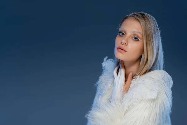 Blonde young woman with winter makeup posing in white faux fur jacket with feathers on dark blue — Stock Photo