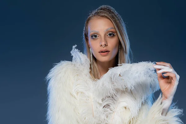 Mujer joven con maquillaje de invierno posando en chaqueta de piel sintética tocando pluma en azul oscuro - foto de stock