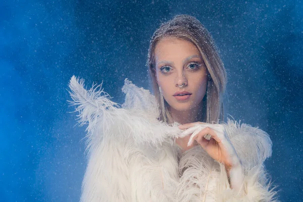 Mujer joven con maquillaje de invierno sosteniendo pluma blanca bajo la nieve que cae en azul oscuro - foto de stock