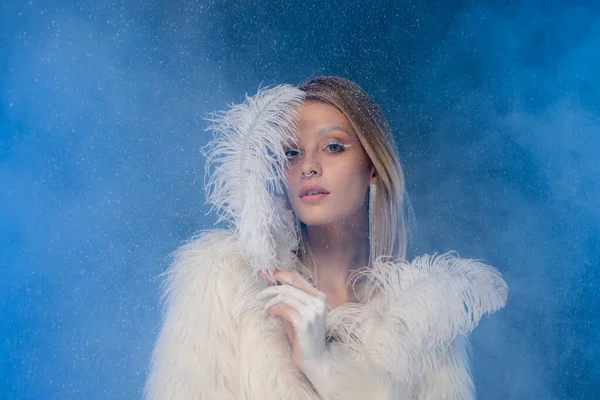 Mujer rubia con maquillaje de invierno en chaqueta de piel sintética sosteniendo pluma blanca bajo la nieve que cae en azul oscuro - foto de stock