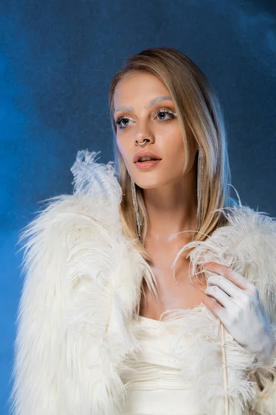 Gepiercte blonde Frau mit Winter-Make-up und gefrorenen Augenbrauen, die Federn auf dunkelblau halten — Stockfoto