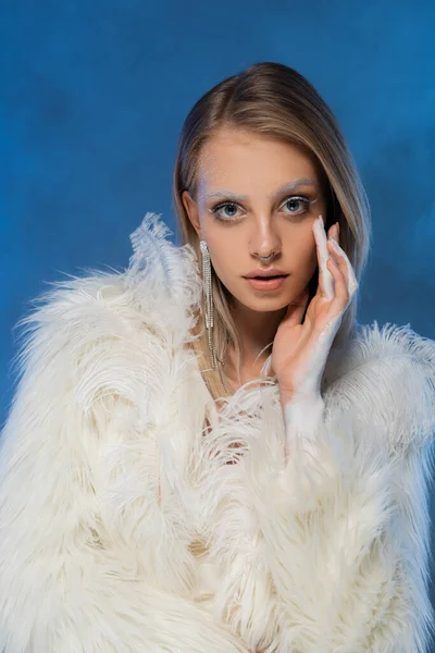 Mujer joven perforada con maquillaje de invierno posando en chaqueta de piel sintética blanca y mirando a la cámara en azul oscuro - foto de stock