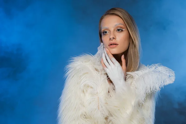 Mujer joven perforada con maquillaje de invierno posando en chaqueta de piel sintética blanca en azul oscuro con humo - foto de stock