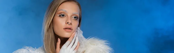 Mujer joven perforada con maquillaje de invierno posando en azul oscuro con humo, pancarta - foto de stock