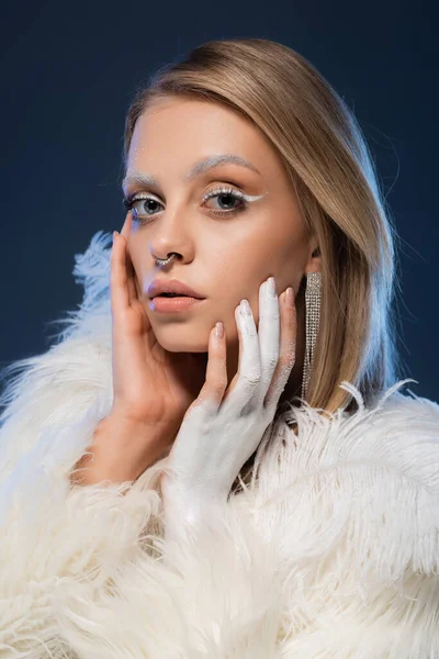 Gepiercte junge Frau mit Winter-Make-up berührt Gesicht auf dunkelblau — Stockfoto