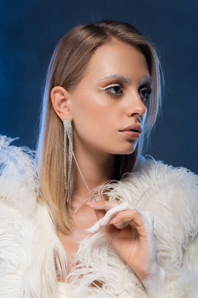 Femme percée avec maquillage d'hiver posant dans une veste blanche en fausse fourrure avec des plumes sur bleu foncé — Photo de stock