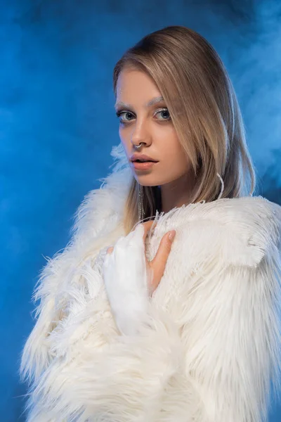 Jeune femme percée avec un maquillage d'hiver posant en veste blanche en fausse fourrure avec des plumes sur bleu avec de la fumée — Photo de stock
