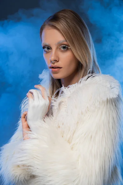 Mujer perforada con maquillaje de invierno posando en chaqueta de piel sintética con plumas en azul con humo - foto de stock