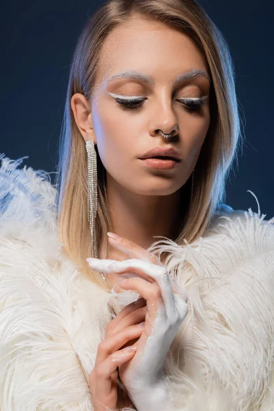 Pierced and blonde woman with winter makeup posing in faux fur jacket with feathers isolated on dark blue — Stock Photo