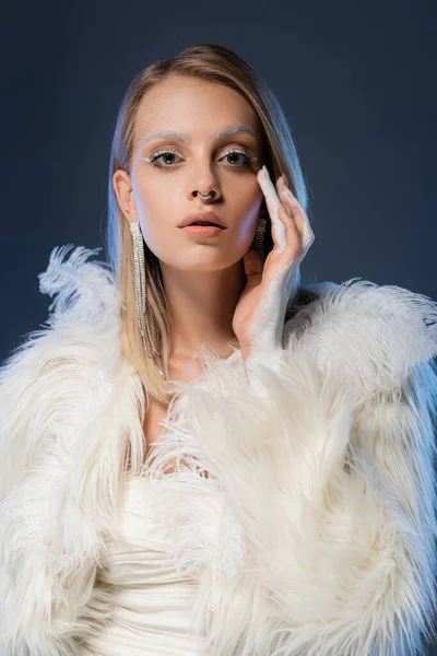 Mujer joven perforada con maquillaje de invierno posando y tocando la cara aislada en azul oscuro - foto de stock