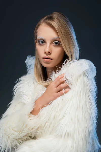 Young blonde woman with winter makeup looking at camera and posing in faux fur jacket isolated on blue — Stock Photo