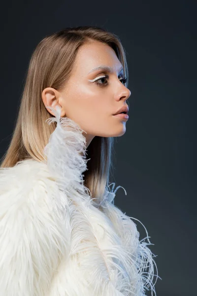 Vista lateral da mulher loira com maquiagem de inverno olhando para longe e posando em casaco de pele falso isolado no azul — Fotografia de Stock