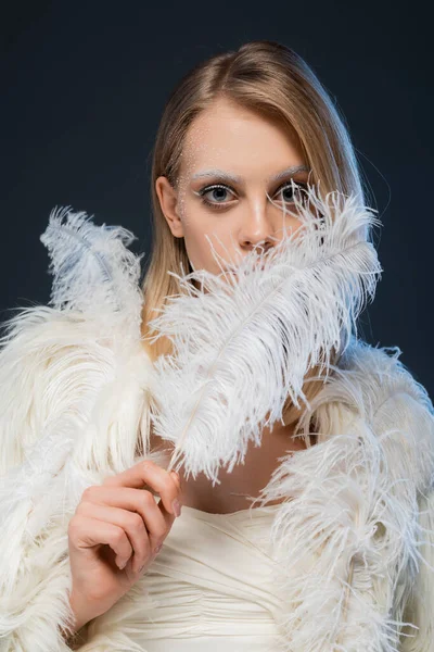 Young blonde woman with winter makeup looking at camera and posing with feather isolated on blue — Stock Photo