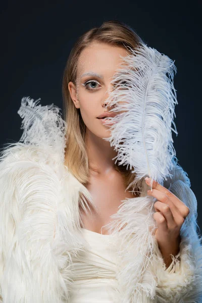 Jeune femme blonde avec maquillage d'hiver regardant la caméra couvrant le visage avec plume isolé sur bleu — Stock Photo