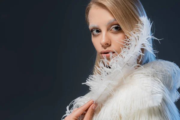 Bonita mujer joven con maquillaje de invierno sosteniendo pluma blanca aislada en azul - foto de stock