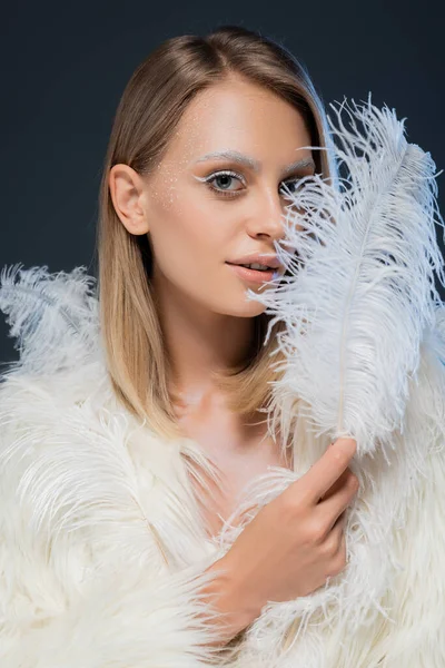 Mujer joven sonriente en chaqueta de piel sintética sosteniendo pluma blanca aislada en azul - foto de stock