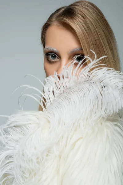 Mujer joven en chaqueta de piel sintética mirando a la cámara a través de una pluma blanca aislada en gris - foto de stock