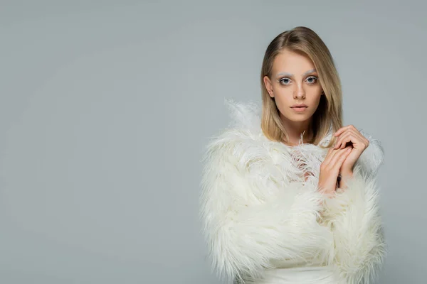 Blonde young woman in faux fur jacket with white feathers looking at camera isolated on grey — Stock Photo