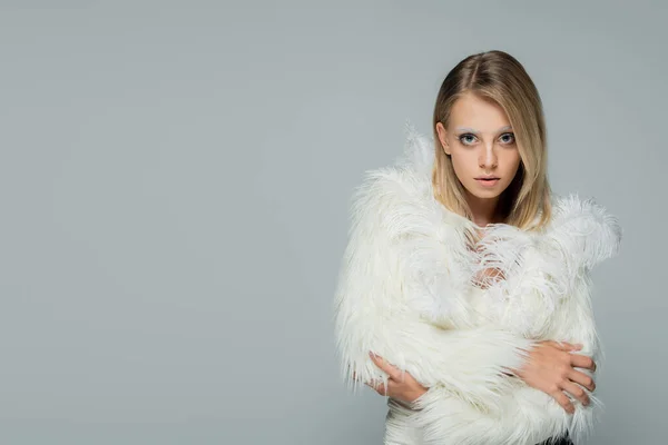 Young woman in stylish faux fur jacket with feathers looking at camera while posing with crossed arms isolated on grey — Stock Photo