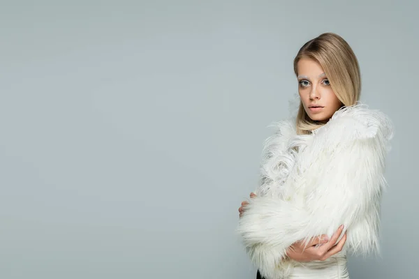 Молодая модель в стильной искусственной меховой куртке с белыми перьями, смотрящая в камеру, позируя изолированно на сером — стоковое фото