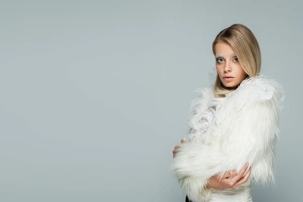 Modelo joven en elegante chaqueta de piel sintética con plumas blancas mirando a la cámara mientras posan aislados en gris - foto de stock