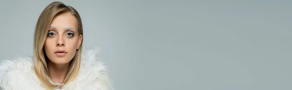 Young model in stylish faux fur jacket with white feathers looking at camera isolated on grey, banner — Stock Photo