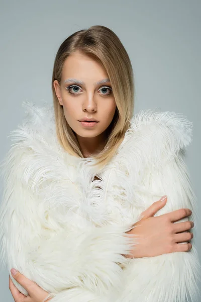 Young model in white faux fur jacket with feathers looking at camera while posing isolated on grey — Stock Photo