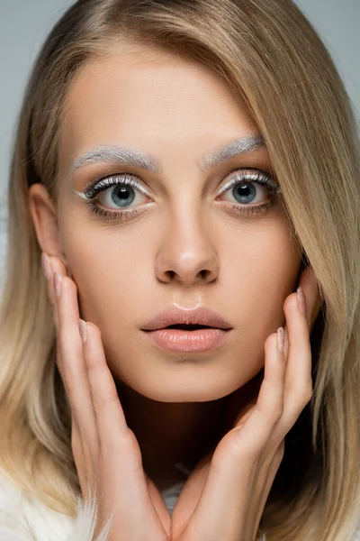 Vista de cerca de modelo joven con maquillaje de invierno y cejas blancas congeladas tocando la cara aislada en gris - foto de stock