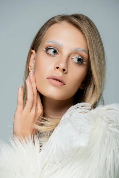 Young model in white faux fur jacket with feathers looking away while posing isolated on grey — Stock Photo