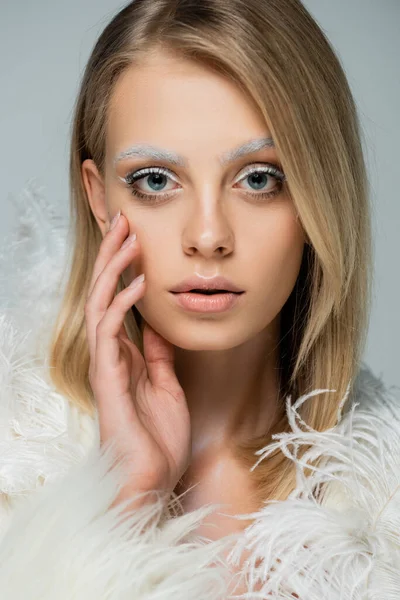 Young model in winter faux fur jacket with feathers looking at camera while touching face isolated on grey — Stock Photo