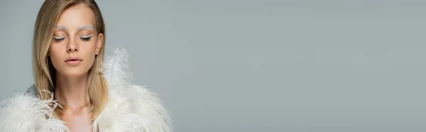 Young woman with winter makeup posing with closed eyes in stylish faux fur jacket with white feathers isolated on grey, banner — Stock Photo