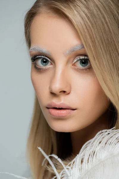 Retrato de jovem mulher com maquiagem de inverno e sobrancelhas brancas congeladas olhando para a câmera perto de penas brancas isoladas em cinza — Fotografia de Stock