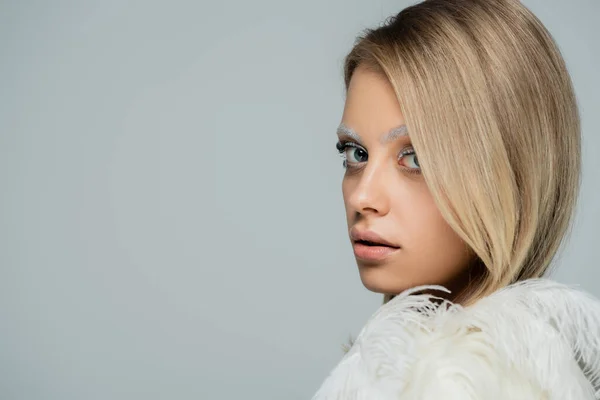 Retrato de jovem mulher com maquiagem de inverno e sobrancelhas brancas olhando para a câmera perto de penas brancas isoladas em cinza — Fotografia de Stock