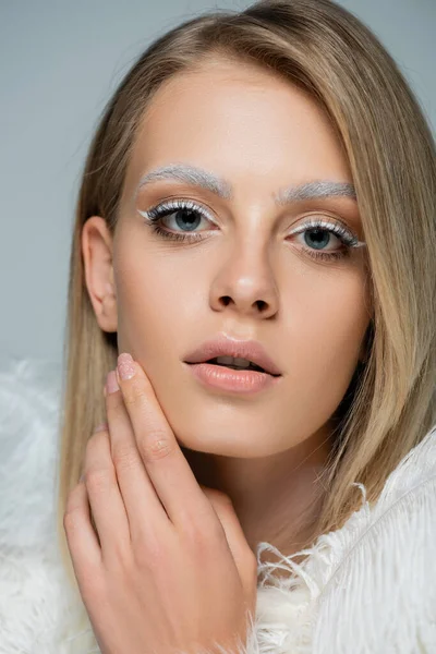 Portrait of young woman with winter makeup and white eyebrows touching face isolated on gray — Stock Photo