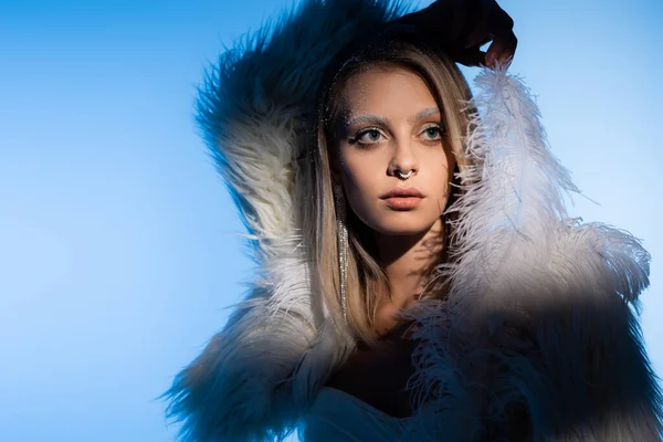 Mulher jovem perfurada com maquiagem de inverno e sobrancelhas brancas posando com pena no azul — Fotografia de Stock