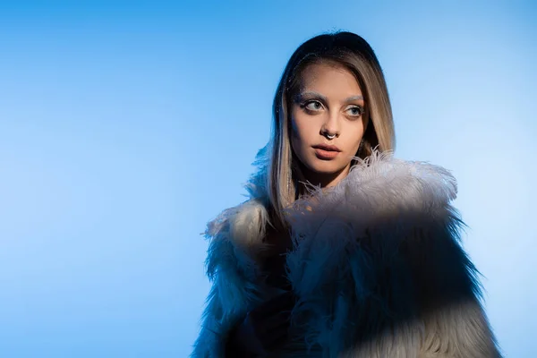 Mujer joven perforada con maquillaje de invierno y cejas blancas posando con pluma sobre fondo azul - foto de stock