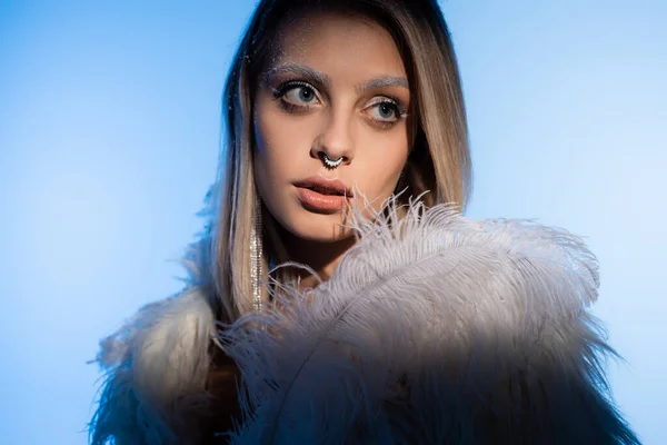 Mujer joven perforada con maquillaje de invierno y cejas blancas posando con pluma mientras mira hacia otro lado en azul - foto de stock