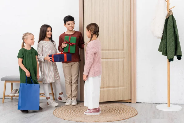 Sorrindo crianças multiétnicas com presentes de pé perto de amigo e porta no corredor em casa — Fotografia de Stock