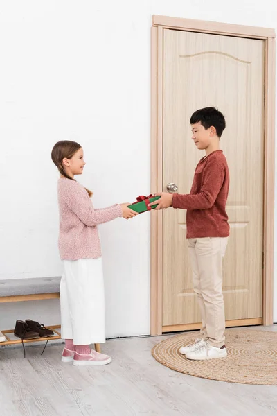Вид сбоку улыбающегося мальчика, делающего подарок другу возле двери дома — стоковое фото
