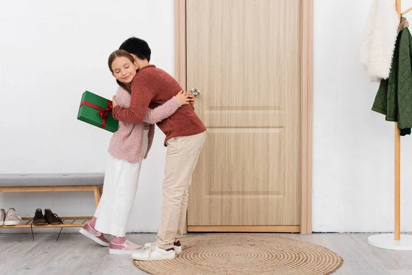 Menino abraçando amigo sorridente e segurando presente perto da porta em casa — Fotografia de Stock