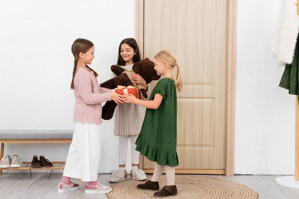 Smiling girls holding soft toy and present near smiling friend at home — Stock Photo