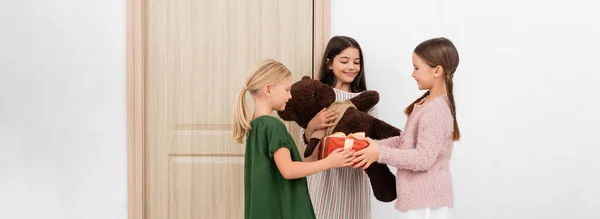 Позитивные девушки дарят мягкие игрушки и подарок возле двери дома, баннер — стоковое фото