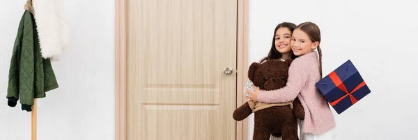 Positive girls with teddy bear and present hugging at home, banner — Stock Photo