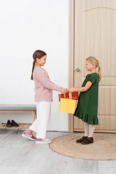 Vue latérale d'un enfant souriant donnant un cadeau et un sac à provisions à un ami à la maison — Photo de stock